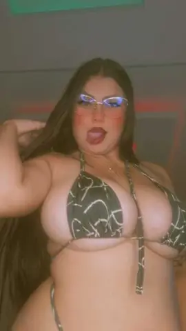 Young woman with long dark hair wearing a patterned bikini top in a soft-lit indoor setting.