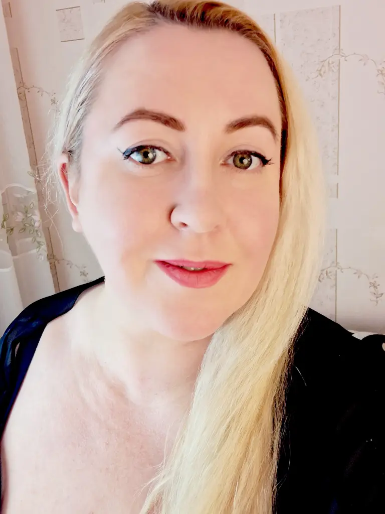 Gingersiya looks at the camera with her blonde hair down, wearing a red top in a room with light patterned wallpaper.