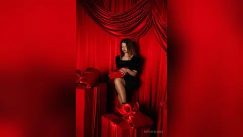 Elegant woman in a black dress sitting amidst red silk drapery and gift boxes in a romantic mood.