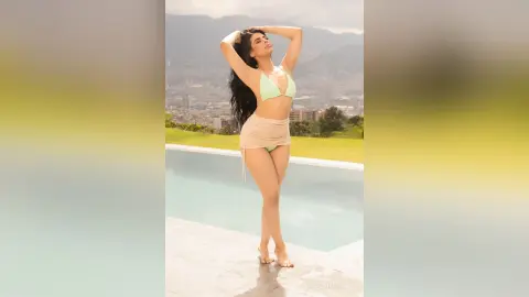 Confident woman in a light green bikini and beige skirt posing by a luxury rooftop pool overlooking a city view.