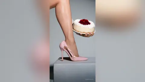 Woman in pink high heels holding a strawberry cream cake against a minimalist grey studio background.