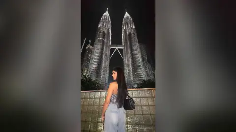 Young woman in light denim jeans posing in front of the illuminated Petronas Twin Towers at night.