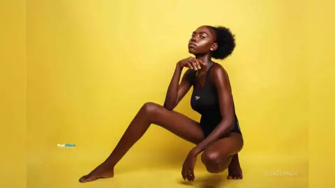 Confident Black woman in a black bodysuit posing against a vibrant yellow studio background.