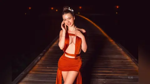 Elie smiles while wearing a low-cut, orange cut-out dress that shows cleavage and her midriff, standing on a wooden walkway at night.