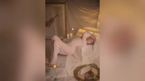 AlanaPearce lies back on white bedding in a white bodysuit, posing near a violin and lit candles in a warm, dim room.