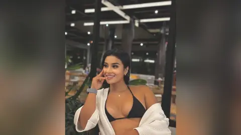 Smiling young woman in a black bikini and white robe relaxing in a modern, dimly lit lounge setting.