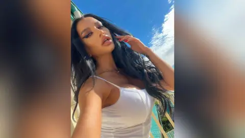 NicoleBella poses in a white tank top with her long dark hair flowing against a clear blue sky.