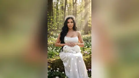 Elegant woman in a white gown and tiara holding a white rabbit in a dreamy, sunlit forest setting.