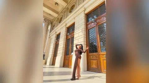 NicoleBella stands against a stone building wall wearing a long brown dress that exposes her legs and midriff.