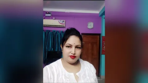 ManishaJi — ManishaJi wears a white top and looks at the camera in a room with purple walls.