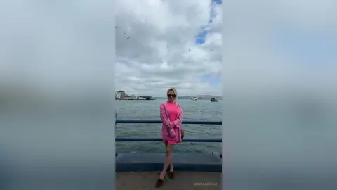 Stylish woman in a pink dress standing on a pier overlooking the ocean under a cloudy sky.