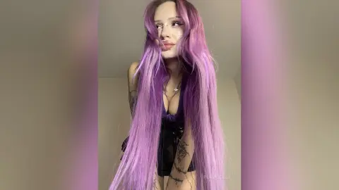 Young woman with long purple hair wearing a black tank top in a soft purple lit indoor setting.