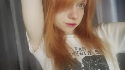 MozelleRozycki poses with her arms raised, wearing a white graphic t-shirt and showing off her long red hair.