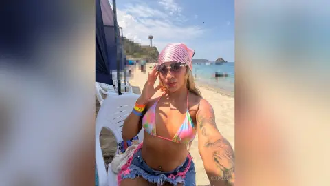 Smiling woman in a colorful bikini and pink headscarf relaxing on a sunny tropical beach.