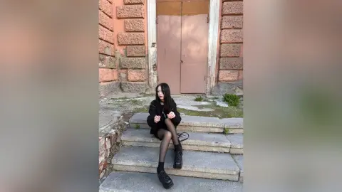 MarieJosePhilip sits on stone steps wearing a black outfit and sheer black tights with black shoes, posing against a large pink door and stone wall.