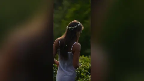 Dreamy woman in a white sundress and flower crown standing in a lush green garden at sunset.