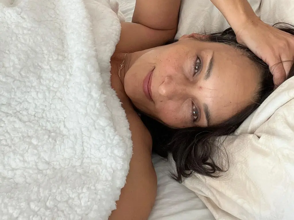 Smiling woman relaxing in bed under a cozy white sherpa blanket in a bright, peaceful bedroom setting.
