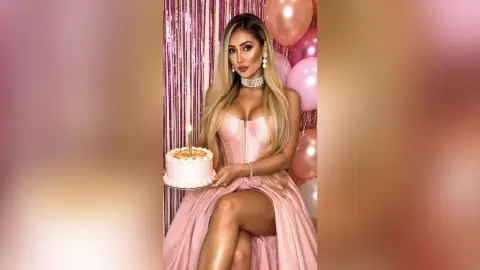 Glamorous woman in a pink satin dress holding a birthday cake amidst pink balloons and shimmering fringe decor.