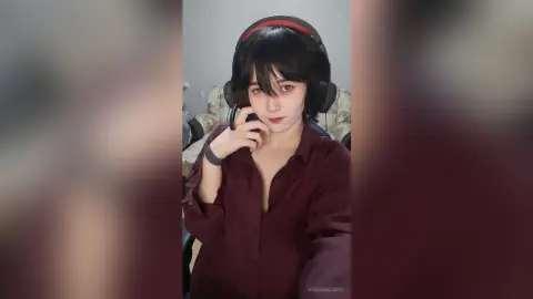 Young woman with short dark hair wearing headphones and a maroon button-down shirt in a cozy indoor setting.