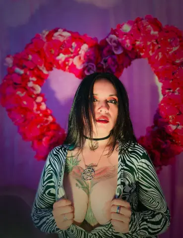 Woman in black and white patterned top posing before a large pink floral heart backdrop in soft lighting.
