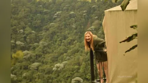 Smiling woman in a dark green sweater relaxing on a balcony overlooking a lush, misty tropical forest.