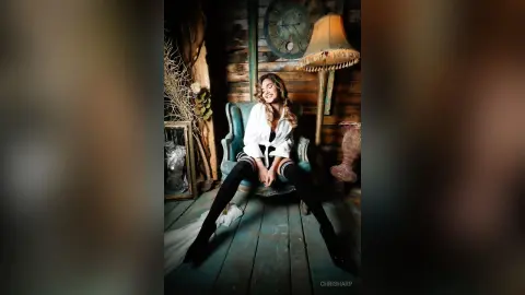 Smiling woman in a white blouse and black leggings sitting in a vintage teal armchair in a rustic room.
