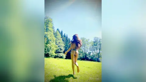 EmaLowee shows her bare back and ass while walking through a sunny, grassy field surrounded by green trees.