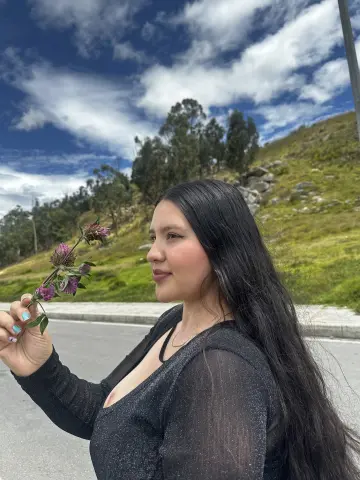 Young woman in a black sparkly top holding a purple wildflower against a sunny mountain landscape.