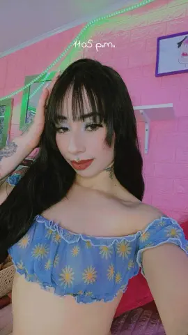 Young woman with dark bangs wearing a blue floral off-the-shoulder crop top in a pink room.