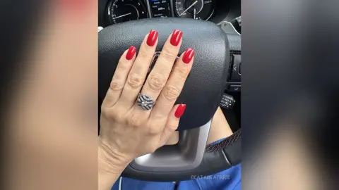 Close-up of a hand with bright red manicured nails and a silver ring resting on a black car steering wheel.