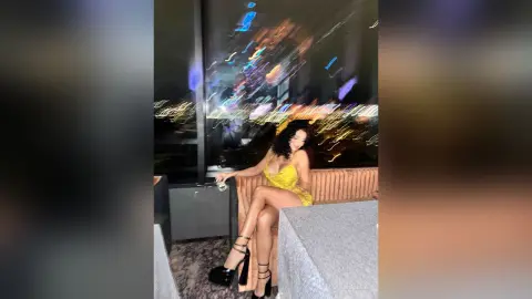 NicoleBella sits on a tan chair wearing a yellow lace dress and black strappy high heels against a blurred city light background.