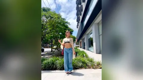 Young woman in a white crochet crop top and blue jeans standing on a sunny city sidewalk near green trees.
