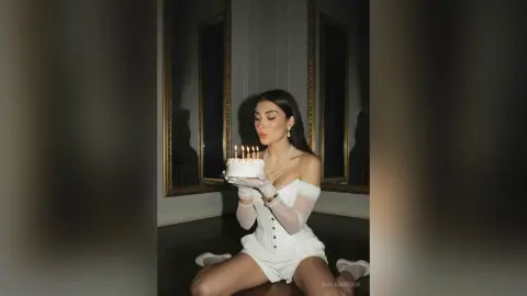 Elegant woman in white corset outfit holding a lit birthday cake in a moody, dimly lit room.