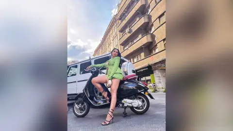 NicoleBella sits on a black scooter wearing a short green outfit and black strappy heels on a city street.