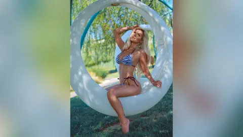 Anita poses in a blue and white striped bikini while sitting on a large white circular swing outdoors.