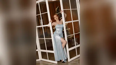 Elegant redhead in a shimmering silver evening gown posing by a window near a festive Christmas tree.