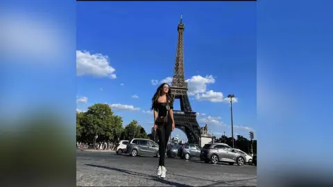 ArianaKroft stands on a cobblestone street in a black bodysuit with long dark hair, posing in front of the Eiffel Tower.