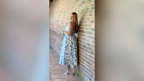 Blonde woman in a blue floral maxi dress standing by a brick wall in soft natural light.