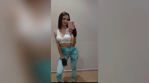 Young woman in white sports bra and teal leggings taking a mirror selfie in a minimalist gym setting.