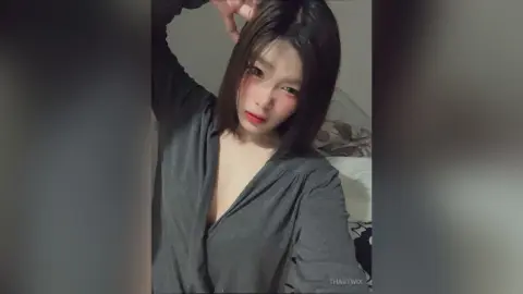 ThaiiTwix poses with her hand in her dark hair, wearing a low-cut grey long-sleeve shirt that shows cleavage.