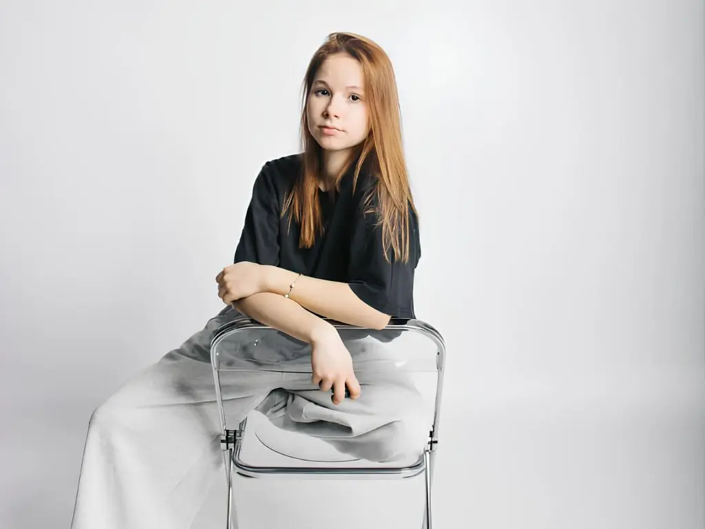LinaForbishh sits on a clear chair wearing a black t-shirt and light grey pants against a white background.