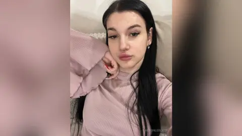 Young woman with long dark hair wearing a pink ribbed long-sleeve top in a cozy indoor setting.