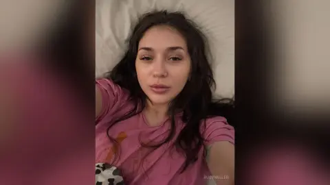 Young woman with long wavy hair wearing a pink t-shirt relaxing on a white pillow in a cozy indoor setting.