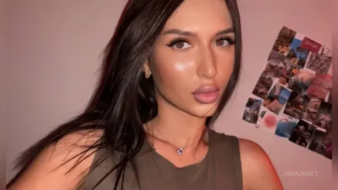 JadaJosey poses for a selfie with long dark hair and a brown tank top against a pink wall.