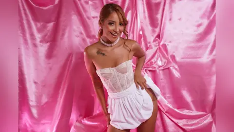 Smiling woman in white lace corset and skirt posing against a glamorous pink satin backdrop.