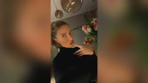 Katia poses with her hand near her mouth, wearing a black turtleneck top in a room with flowers.