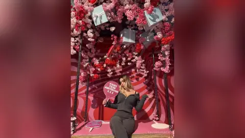ValerieGracy poses in a black long-sleeved top against a pink floral backdrop with red and pink flowers hanging above her.