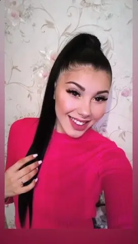 Smiling young woman with long dark ponytail wearing a bright pink sweater against a floral wallpaper background.