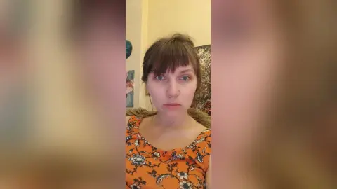 StacyMalvin looks at the camera with brown hair and bangs, wearing an orange floral print tank top against a plain wall background.