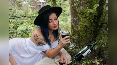 Woman with blue hair in a white dress enjoying red wine by a mossy tree in a lush garden setting.